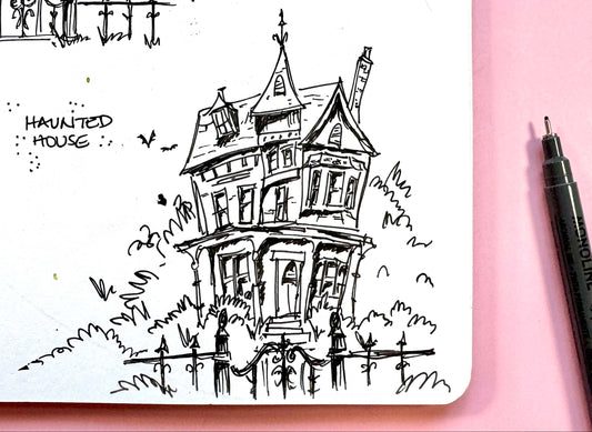 A black ink sketch of a haunted house in a small notebook on a pink background, with a black pen visible on the right side.